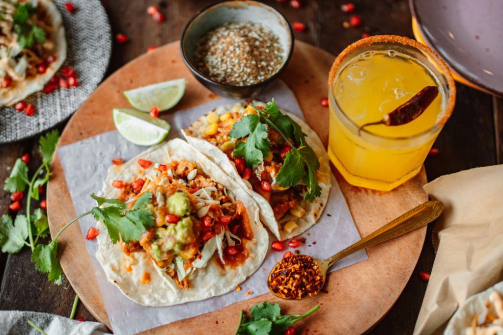 Zbliżenie na catering wegetariański serwujący tacos z salsą, świeżą kolendrą, ziarnami granatu i guacamole, podane obok drinka na drewnianej tacy.