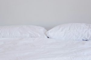 Na zdjęciu przedstawiono łóżko z białą pościelą i dwiema dużymi poduszkami opartymi o jasną ścianę.