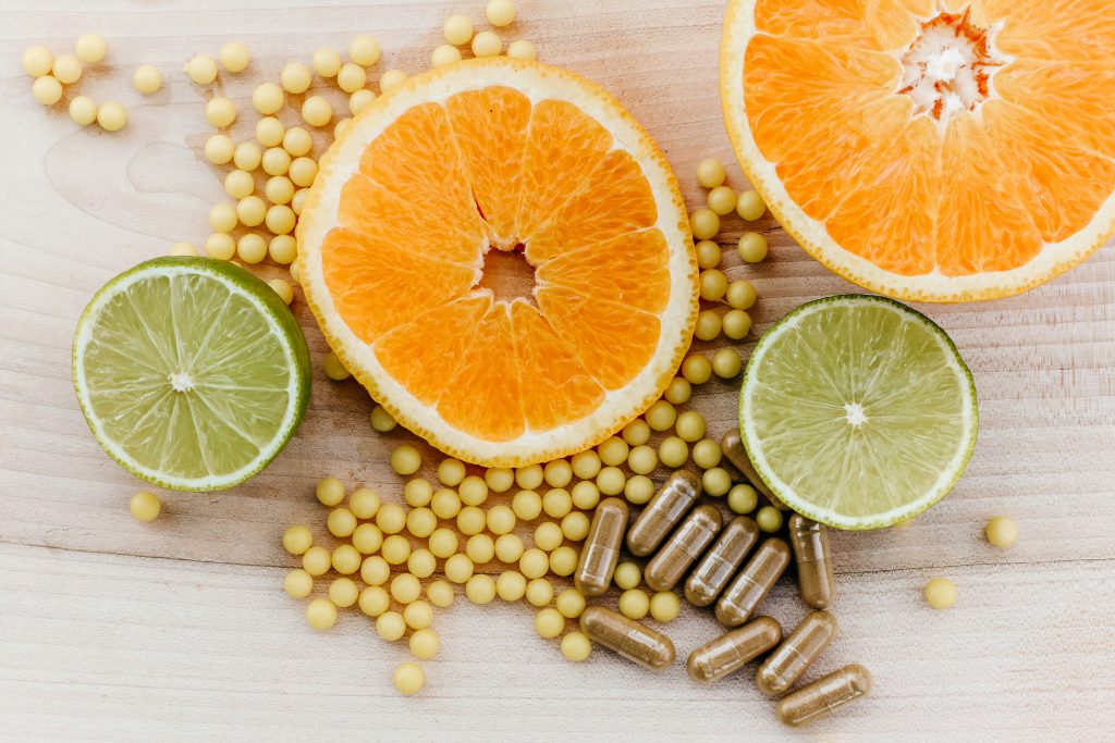 zdjęcie przedstawia kompozycję złożoną z przekrojonych owoców cytrusowych – pomarańczy i limonki – ułożonych na jasnej, drewnianej powierzchni, wokół nich rozsypane są małe, żółte kulki oraz brązowe kapsułki z proszkiem w środku